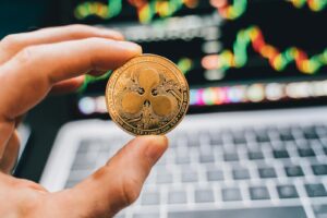 A close-up view of a hand holding a Ripple cryptocurrency coin with a blurred computer in the background.